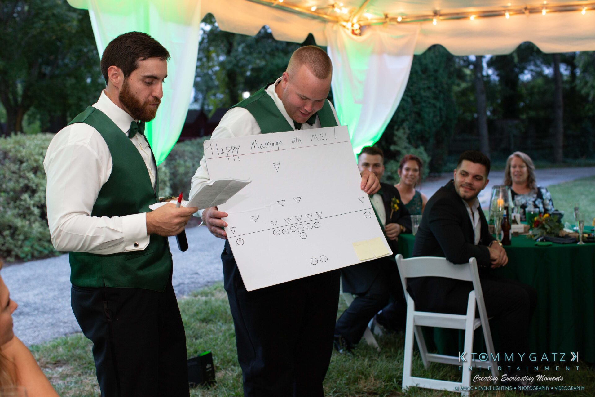 Two men in vests give a wedding speech outdoors, holding a presentation board. Guests sit at tables under a tent with green lighting.