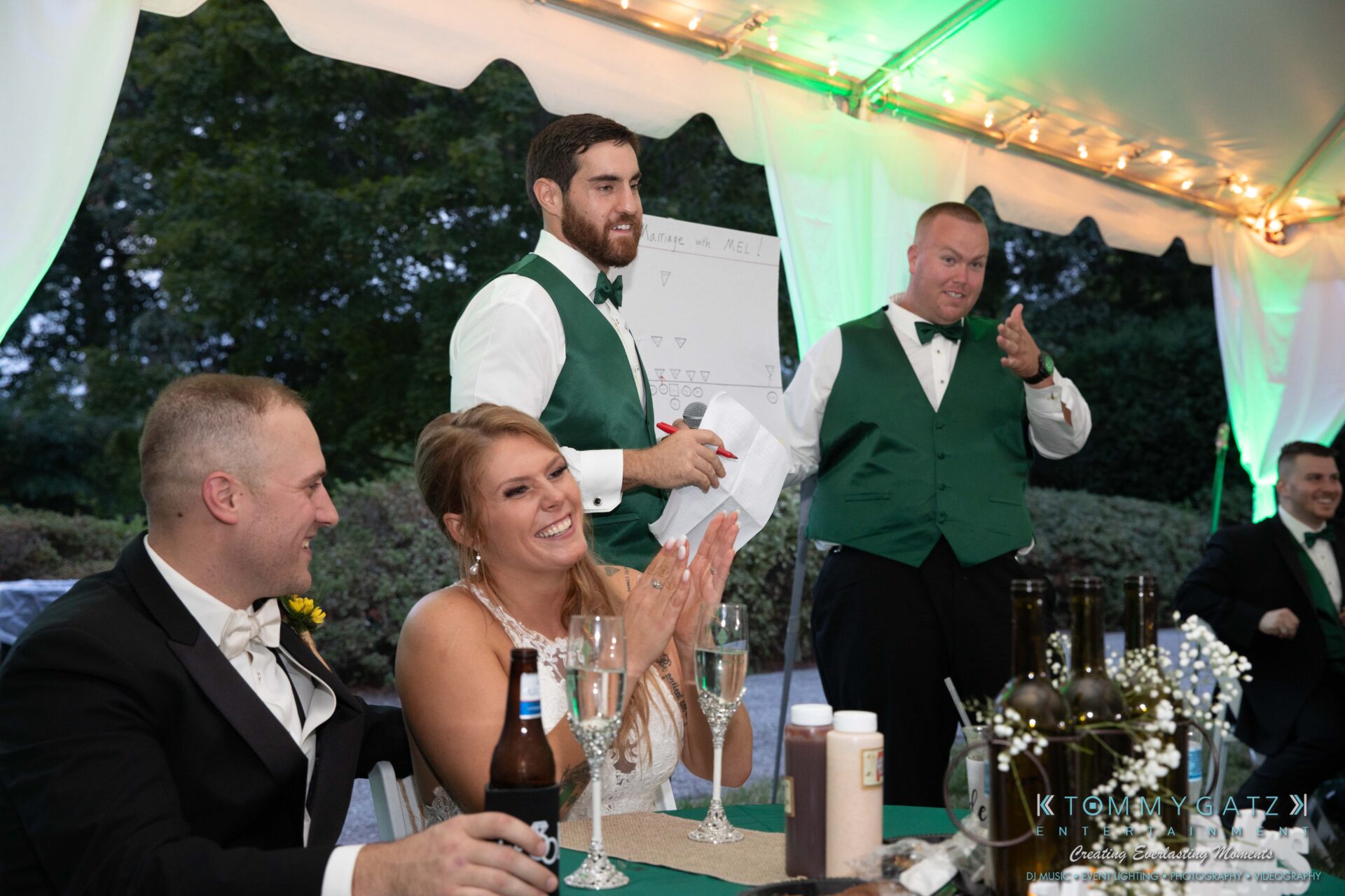 Proctor-Wedding-HIGHLIGHTS-091419-92 - TOMMY GATZ ENTERTAINMENT A bride and groom at a table with guests. One guest is standing, holding a microphone and speaking. The setting is an outdoor tented event with a board and bottles visible on the table.