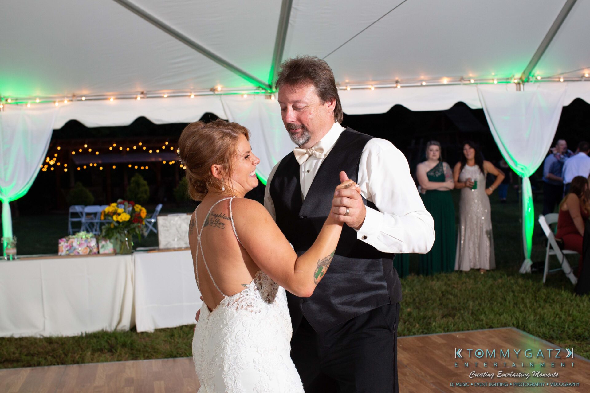 Proctor-Wedding-HIGHLIGHTS-091419-95 - TOMMY GATZ ENTERTAINMENT A couple dances under a white tent with green lighting. The woman wears a white dress, the man a black vest and bow tie. Guests and a decorated table are in the background.