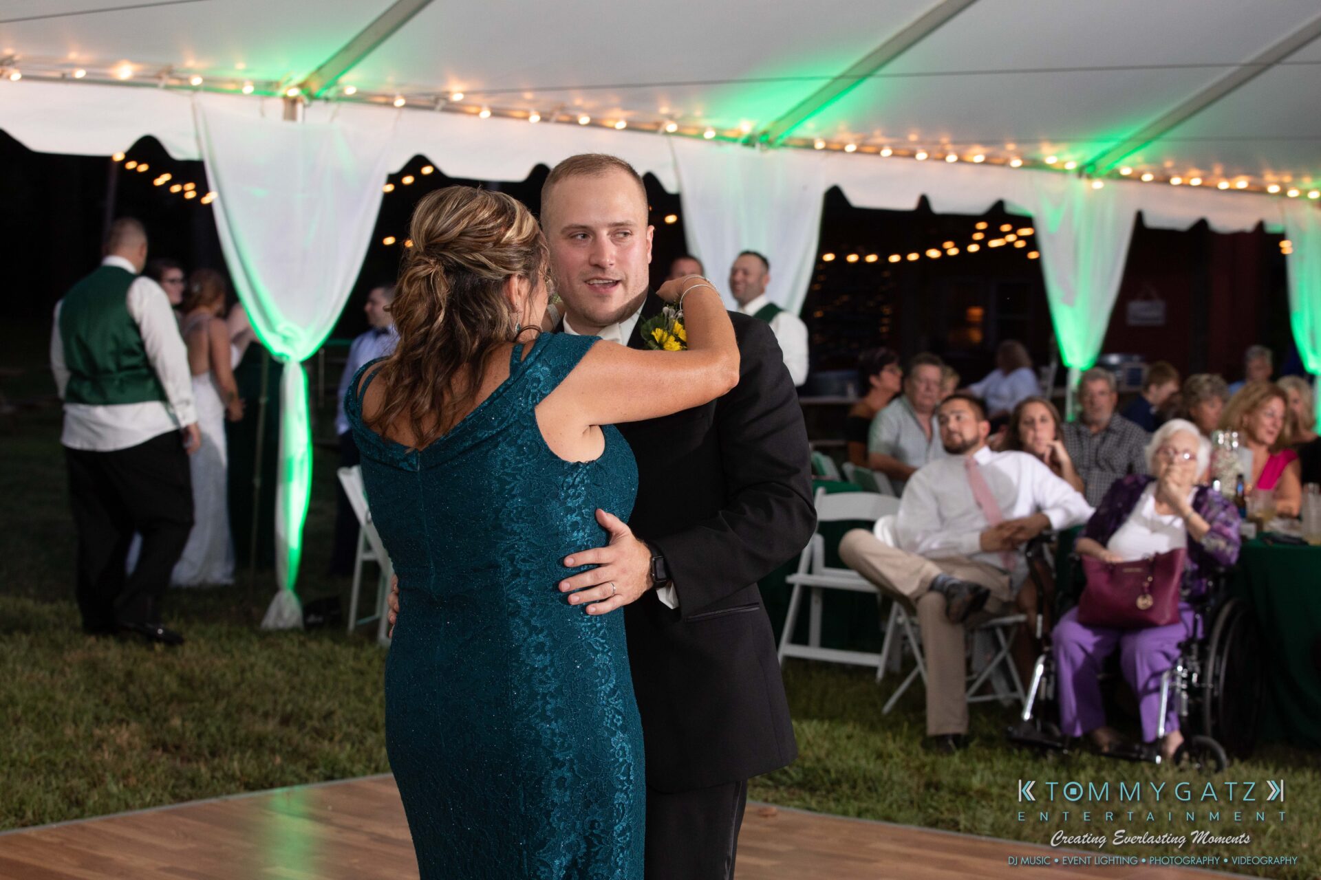 Proctor-Wedding-HIGHLIGHTS-091419-97 - TOMMY GATZ ENTERTAINMENT A man in formal wear dances with a woman in a green dress under a tent at a wedding reception. Guests are seated and watching.