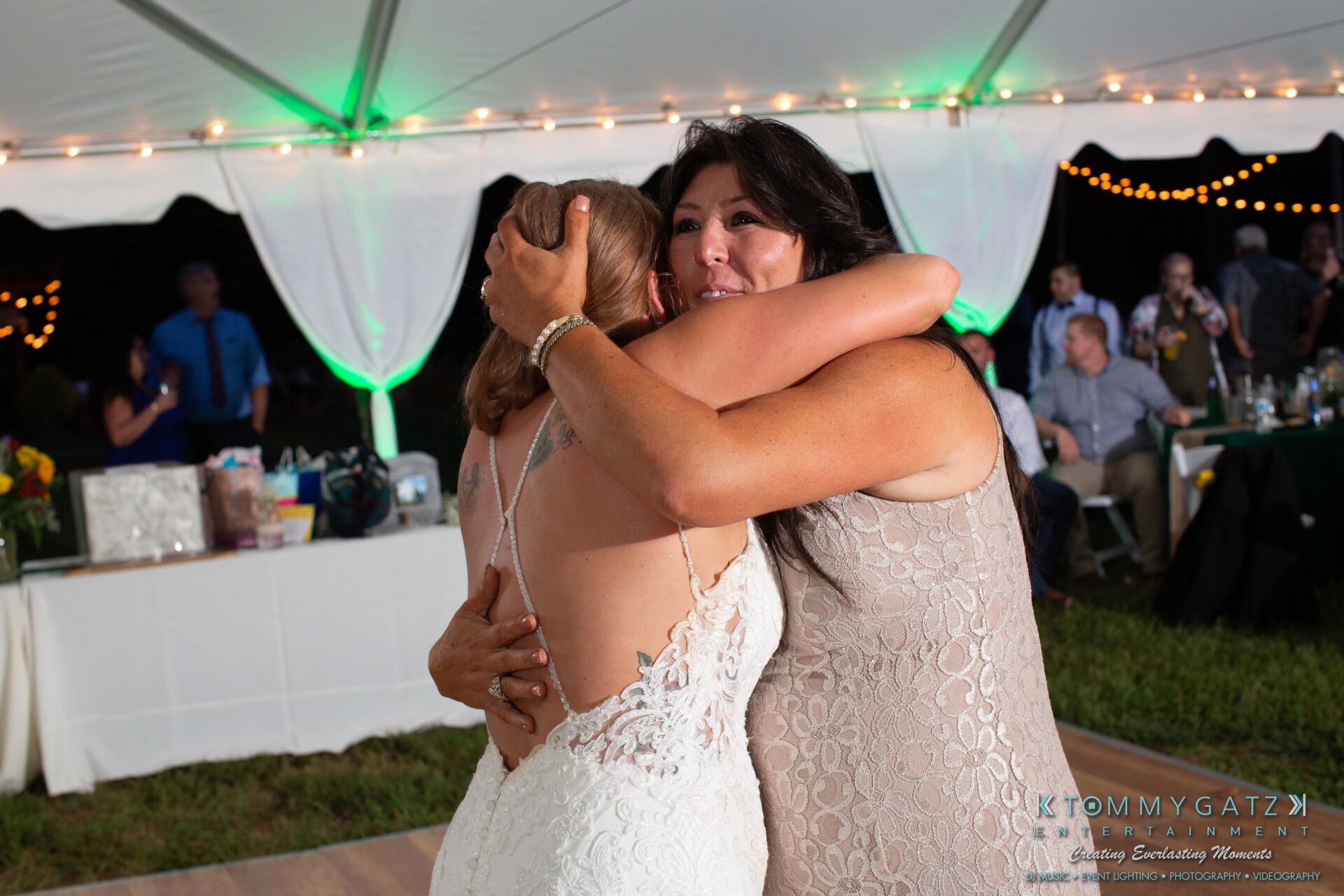 Proctor-Wedding-HIGHLIGHTS-091419-99 - TOMMY GATZ ENTERTAINMENT Two women in formal dresses embrace under a tent with string lights. Guests are seated at tables in the background.