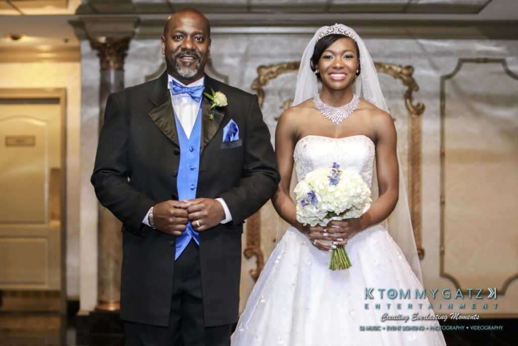 A bride in a white gown and tiara stands beside a man in a tuxedo with a blue vest and bow tie, holding a bouquet.
