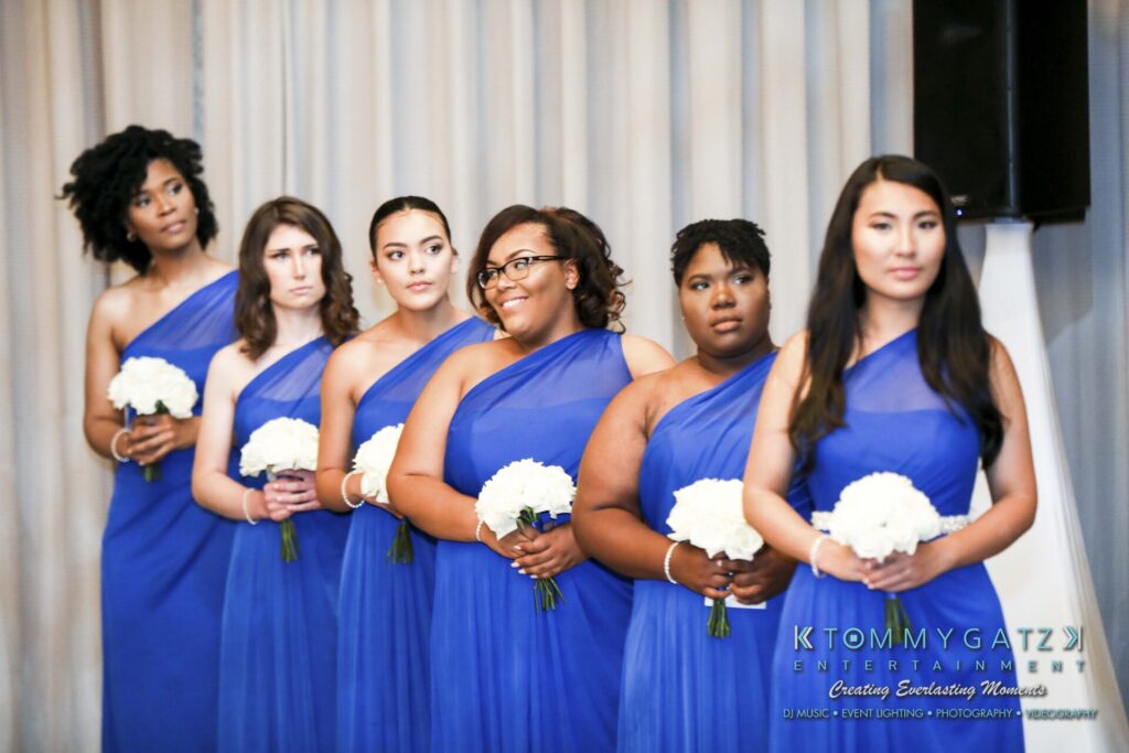 Six bridesmaids in matching blue dresses hold white bouquets, standing in a line against a light-colored curtain backdrop.