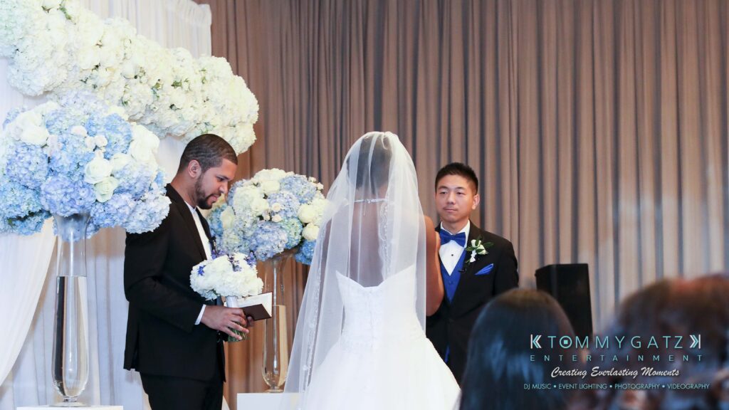 Bride and groom stand facing each other during wedding ceremony, with officiant holding a book nearby. White floral arrangements decorate the background.