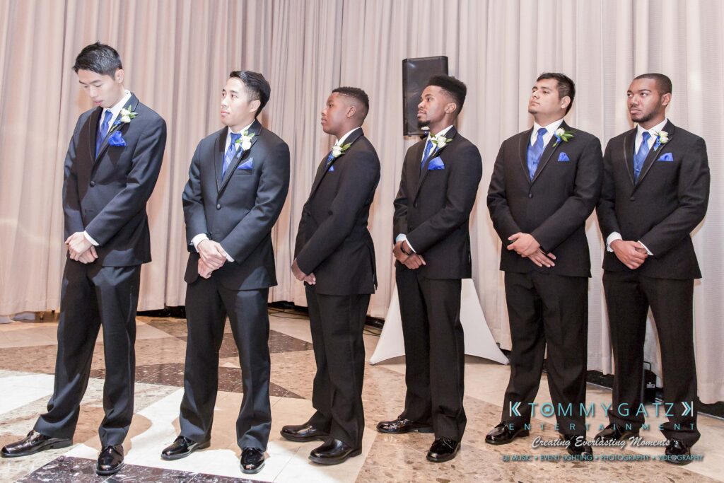 Six groomsmen in black suits with blue accents stand in a line indoors, in front of light-colored curtains.