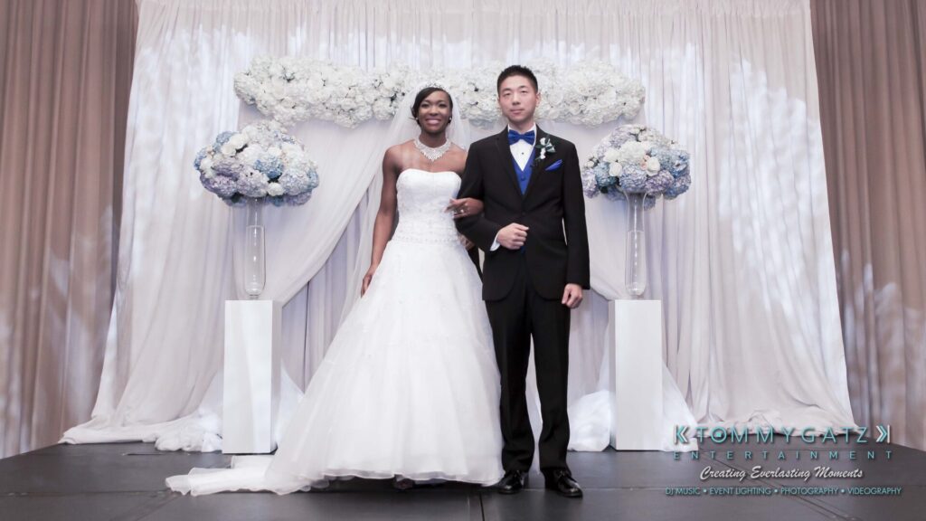 Bride in a white gown and groom in a black tuxedo stand on a stage with floral arrangements behind them.