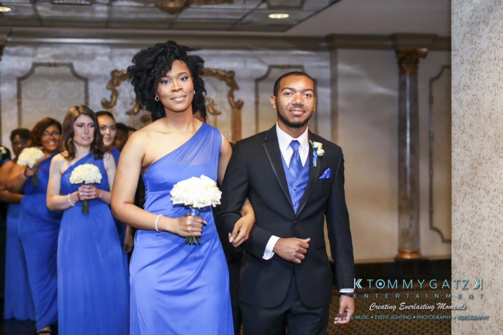 A group of people in formal attire walk in pairs down an aisle. The women wear blue dresses and hold bouquets, while the men wear black suits with blue vests and ties.