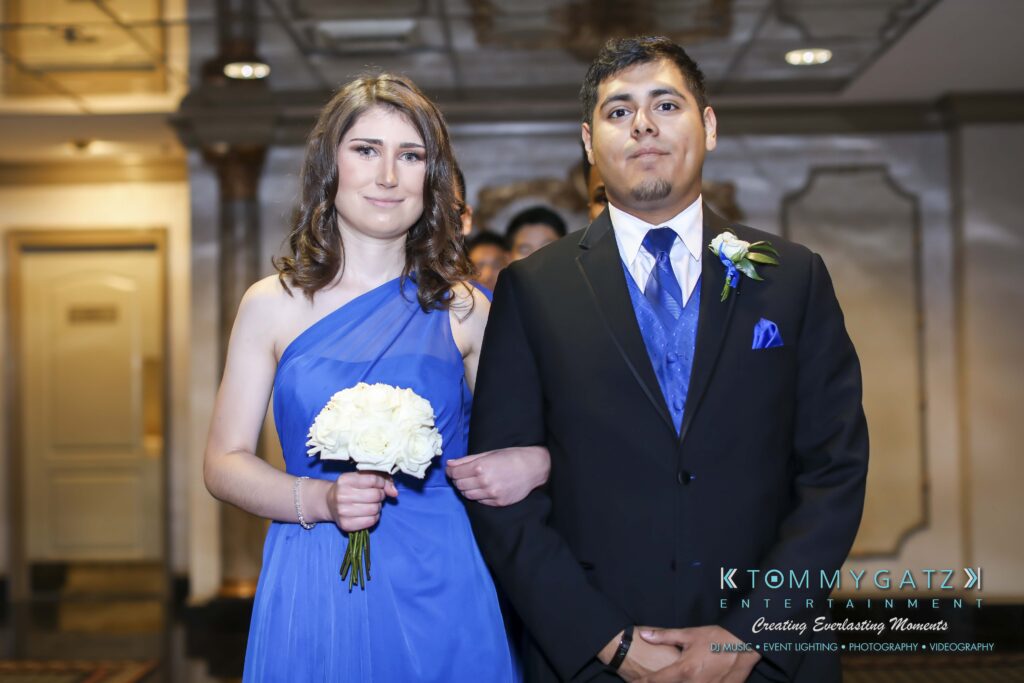 A couple stands together at a formal event. The woman is in a blue dress holding white flowers, and the man is in a dark suit with a blue tie.