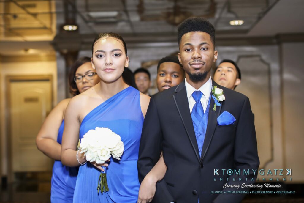 A group of people in formal attire at an event. A man and woman, both dressed in blue, are linked arm-in-arm. The woman holds a bouquet of white flowers. Others are visible in the background.