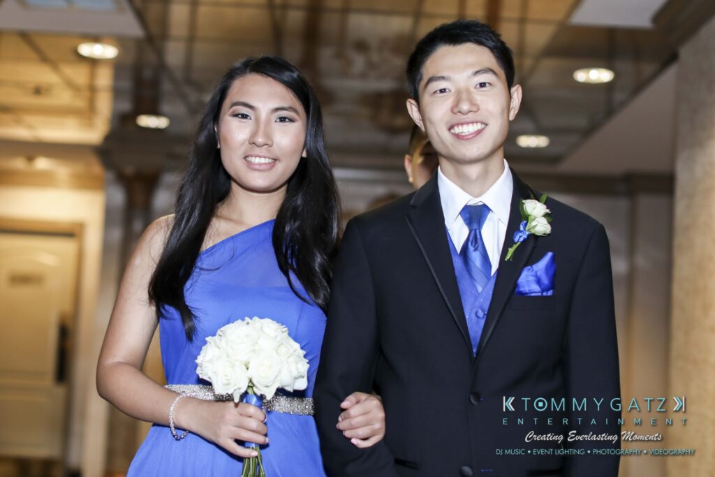 A young couple in formal attire smiles, with the woman holding a bouquet of white flowers.