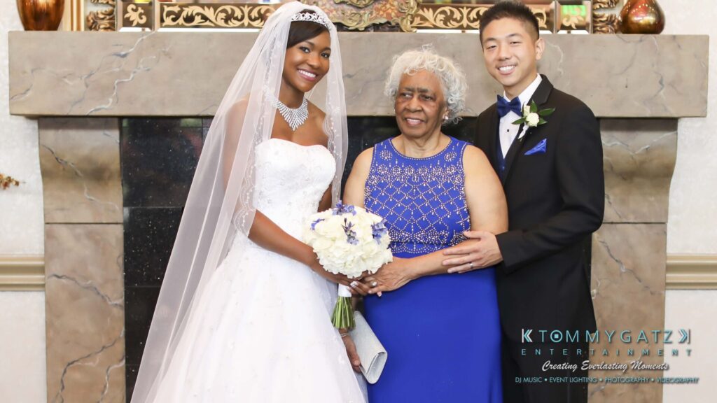 A bride, an older woman, and a groom pose together in formal attire at a wedding. The bride holds a bouquet.
