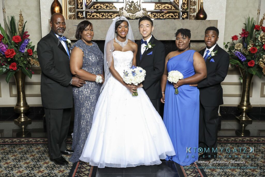 A bride and groom pose with four family members. Everyone is dressed formally, with the bride in a white gown and others in suits and dresses.