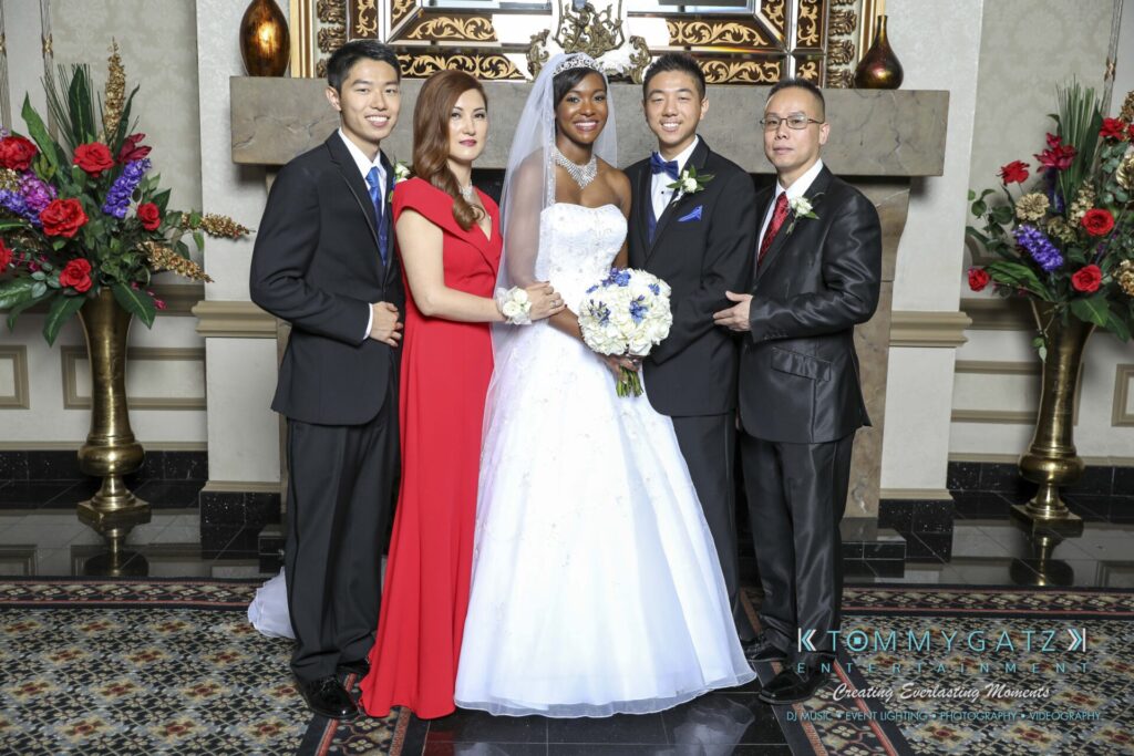 A bride and groom stand with three family members in formal attire, posing for a photo in an ornate room with flowers and patterned flooring.