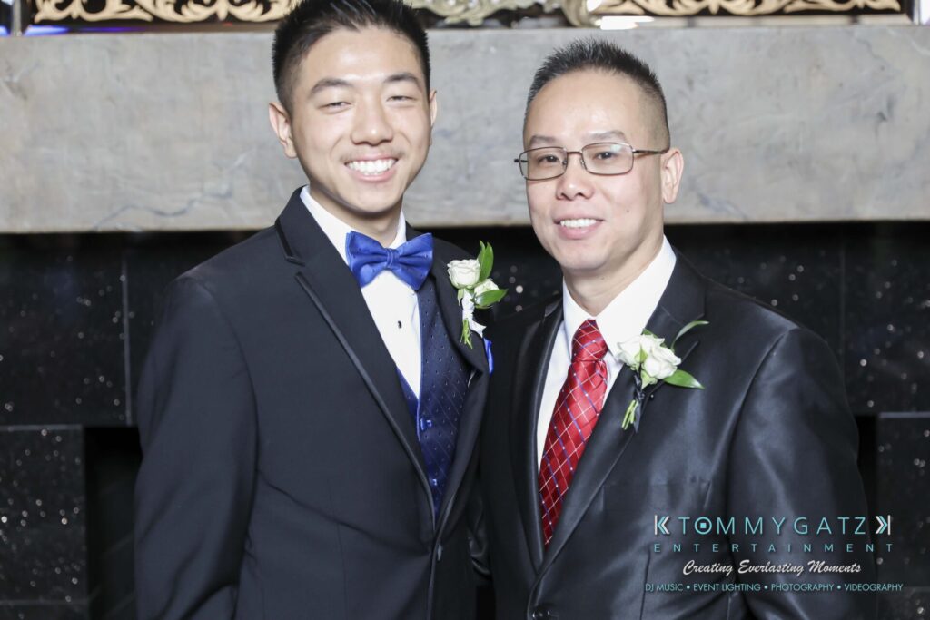 Two people in suits, wearing boutonnières, stand side by side smiling. The backdrop is a dark, elegant setting with a decorative element above.