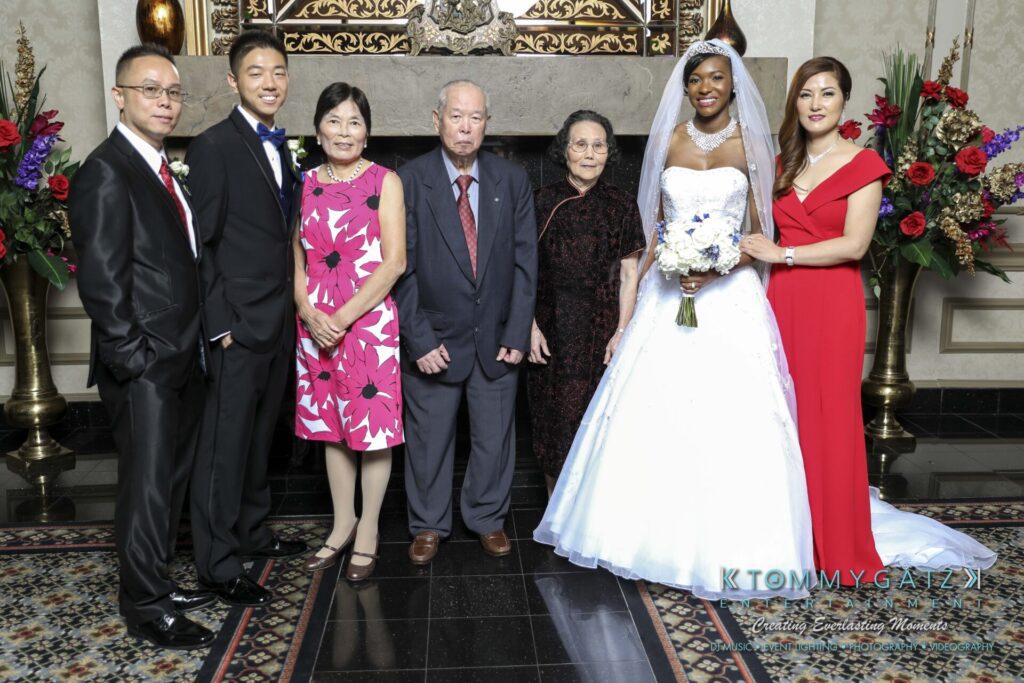 A bride in a white gown stands with a groom and five other people in formal attire in front of a fireplace with floral arrangements.