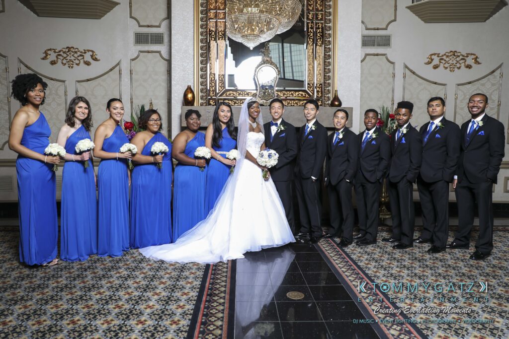A bridal party posing indoors. The bride and groom stand at the center, surrounded by bridesmaids in blue dresses and groomsmen in black suits with blue ties.