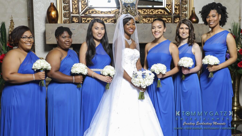 A bride in a white gown stands with six bridesmaids in blue dresses, all holding bouquets, in front of a decorative mirror.