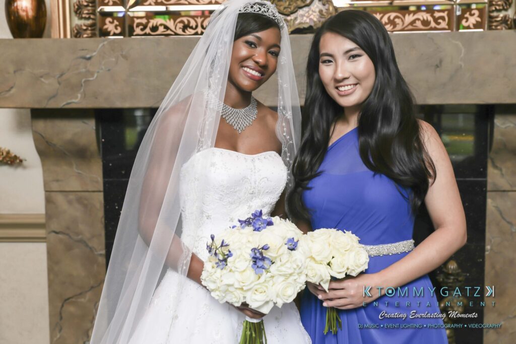 A bride in a white dress and veil stands next to a woman in a blue dress, both holding white flower bouquets, in front of a fireplace.