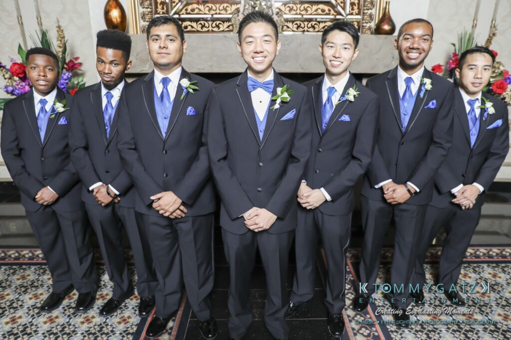 Seven men in suits and blue vests stand in a line, smiling at the camera. There's a decorative fireplace and floral arrangement in the background.