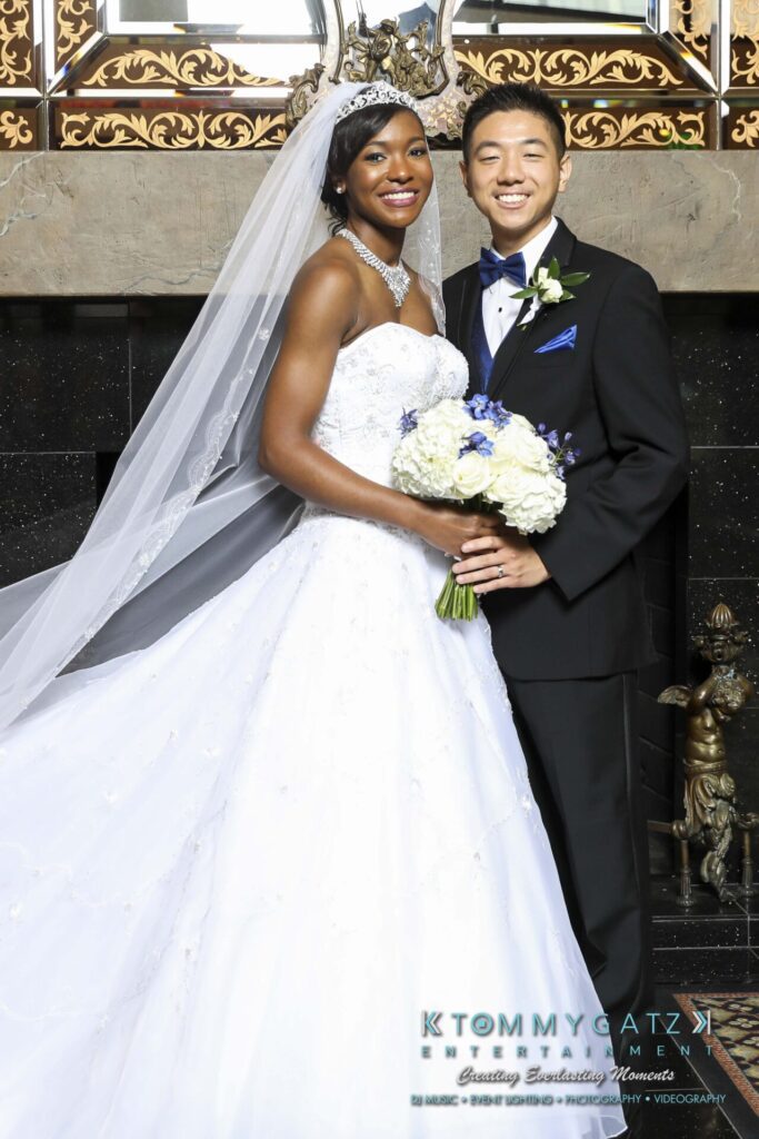 A bride in a white gown and tiara stands beside a groom in a black tuxedo, holding a bouquet of white and blue flowers, in front of an ornate backdrop.