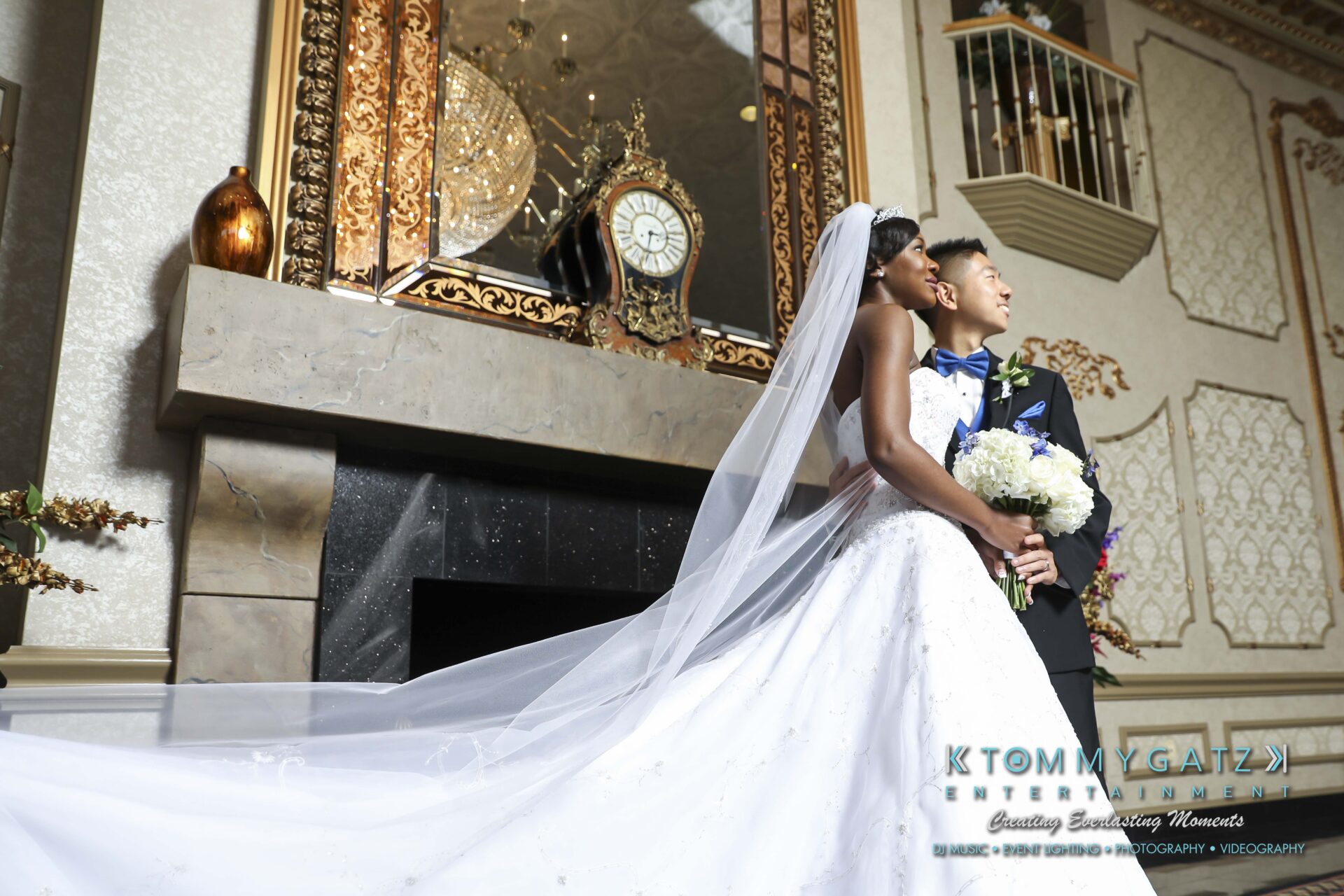 A bride and groom stand together indoors, dressed in wedding attire. The bride holds a bouquet and wears a long veil. They are near a fireplace with decorative mirrors and a clock above.