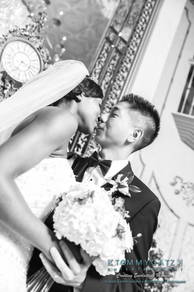 Raneka-Michael-FORMALS-Low-Res-WM-141 - TOMMY GATZ ENTERTAINMENT A bride and groom share a kiss in a black and white photo. The bride holds a bouquet, and both are dressed in formal wedding attire. Ornate decorations are visible in the background.