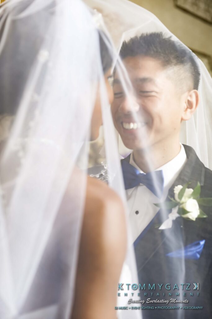 A bride and groom smile at each other under a veil. The groom wears a tuxedo with a blue bow tie and boutonniere. The bride's face is partially visible.