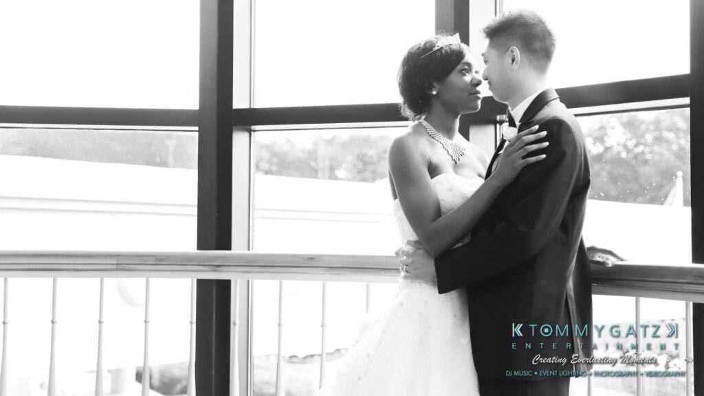 A couple in formal attire embraces in front of large windows, with a view of trees outside.