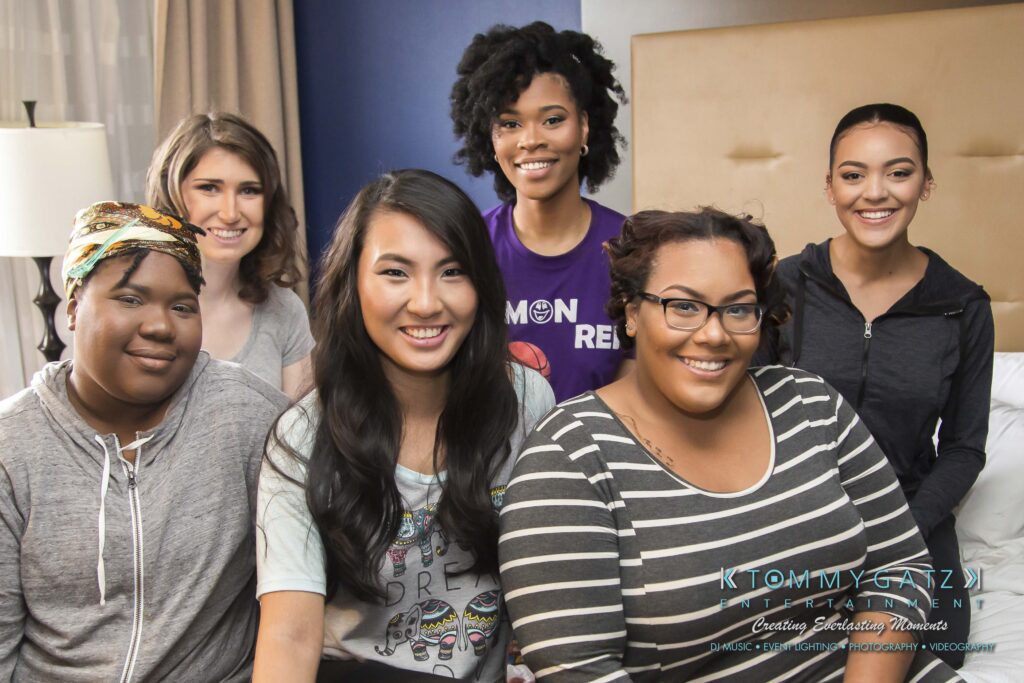 Six people smiling, seated indoors in a casual setting. Some are wearing casual tops with graphics. A bed and curtains are visible in the background.