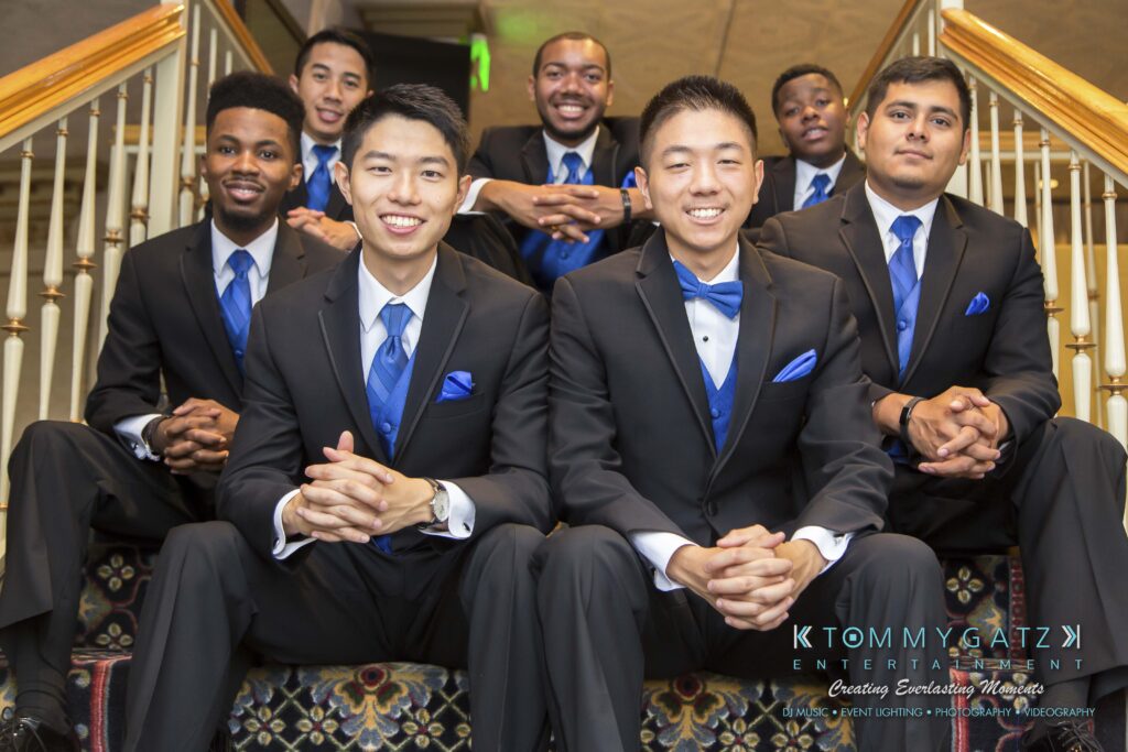 Raneka-Michael-PREP-Low-Res-WM-11 - TOMMY GATZ ENTERTAINMENT A group of seven men in black suits with blue ties sit on a staircase, smiling at the camera.