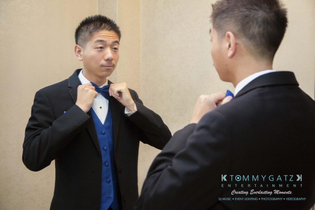 A man in a suit adjusts his blue bow tie while looking in a mirror.