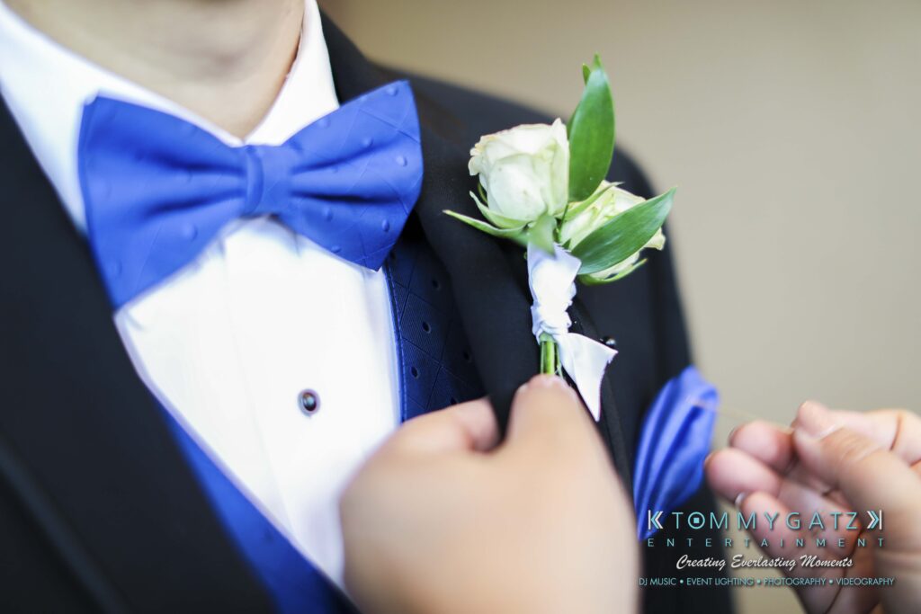 Person in a black suit and blue bow tie getting a white boutonniere pinned to their lapel by another hand. Logo in the corner reads'Tommy Gatz Entertainment.'.