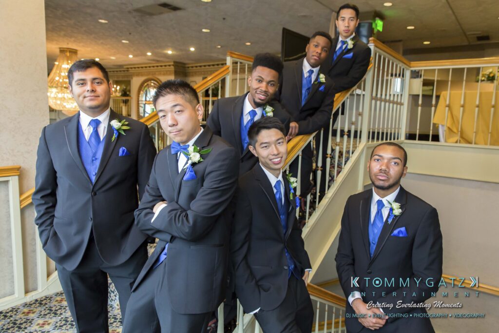 A group of seven men in black suits with blue vests and ties pose on a staircase indoors.