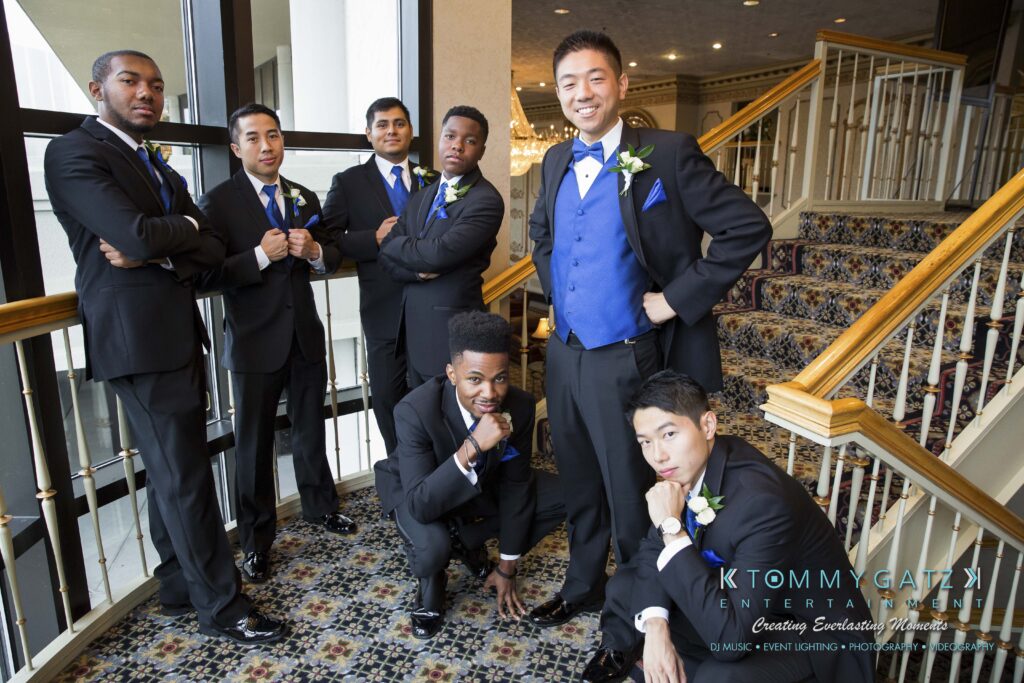 A group of groomsmen in black suits with blue vests and ties pose on a staircase, some standing and some kneeling, with hands clasped or on chins.