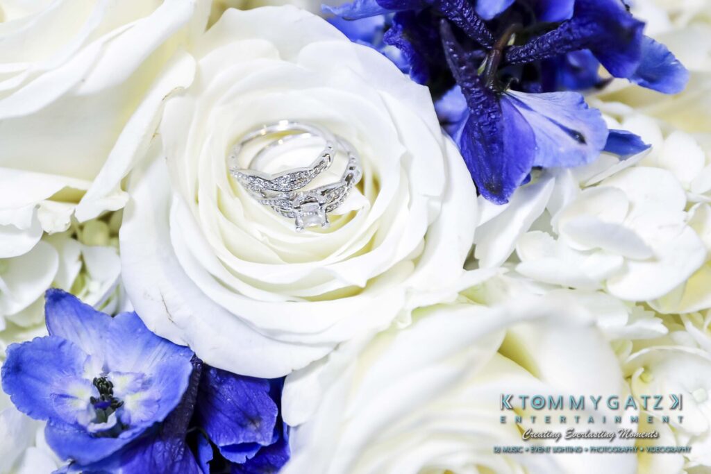 Close-up of wedding rings on a white rose surrounded by white and purple flowers.
