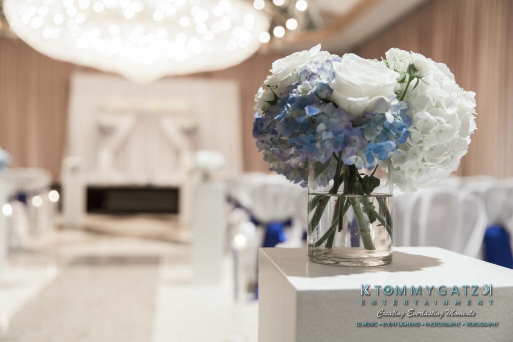 Glass vase with blue and white hydrangeas on a pedestal in a decorated room, featuring draped fabric and a chandelier.