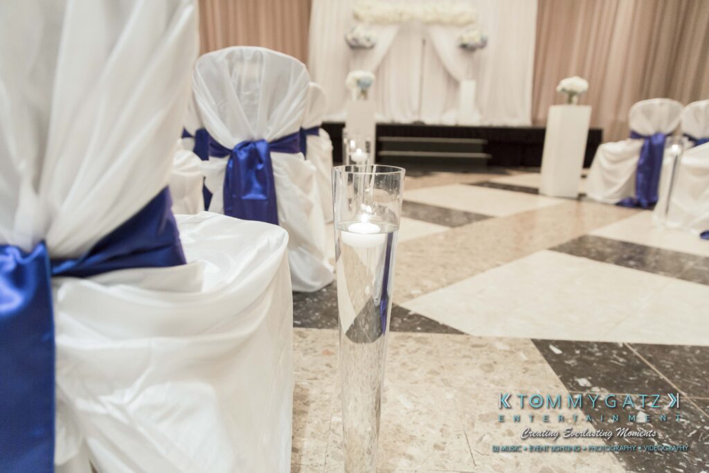 Wedding ceremony setup with rows of white chairs adorned with blue sashes. A single lit candle in a glass holder is in the foreground on a black and white tiled floor.
