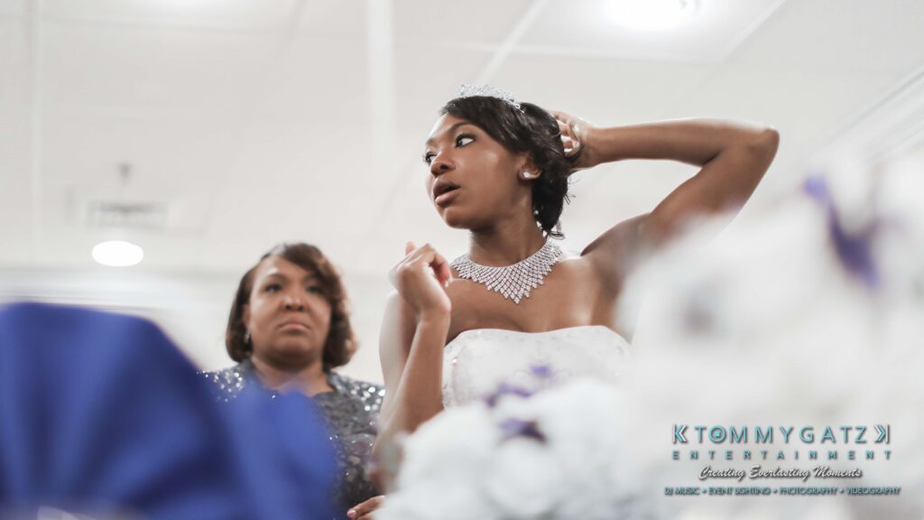 A bride wearing a white gown and jeweled necklace adjusts her hair. A woman stands nearby, watching. A blurred blue shape is in the foreground.