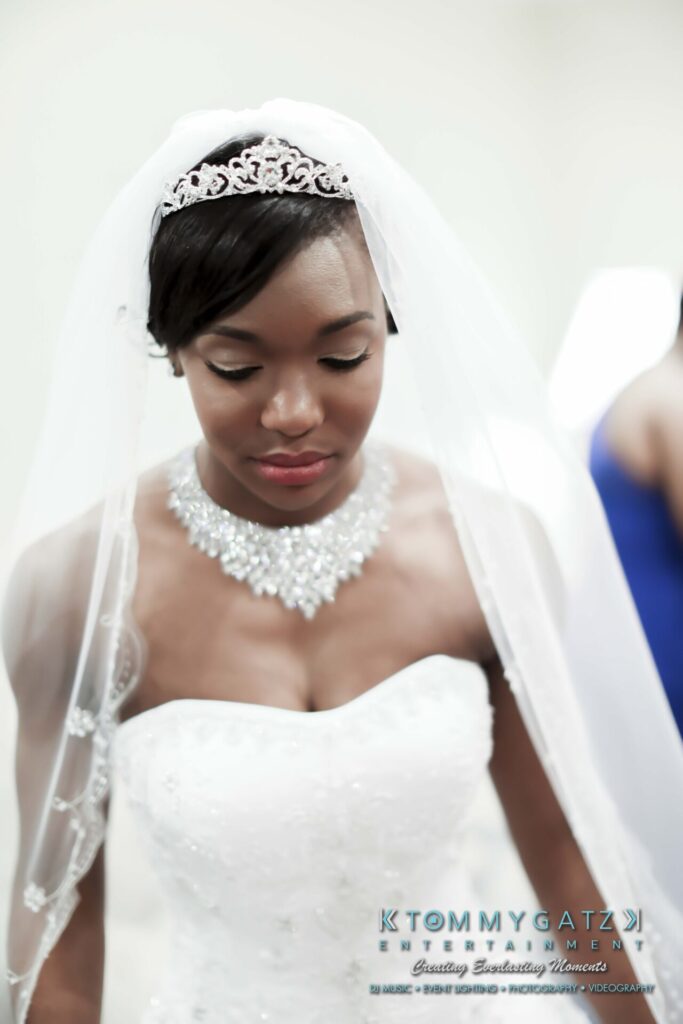 Raneka-Michael-PREP-Low-Res-WM-41 - TOMMY GATZ ENTERTAINMENT Bride in a white strapless gown with an ornate necklace and tiara, looking downward. Veil drapes over her shoulders.