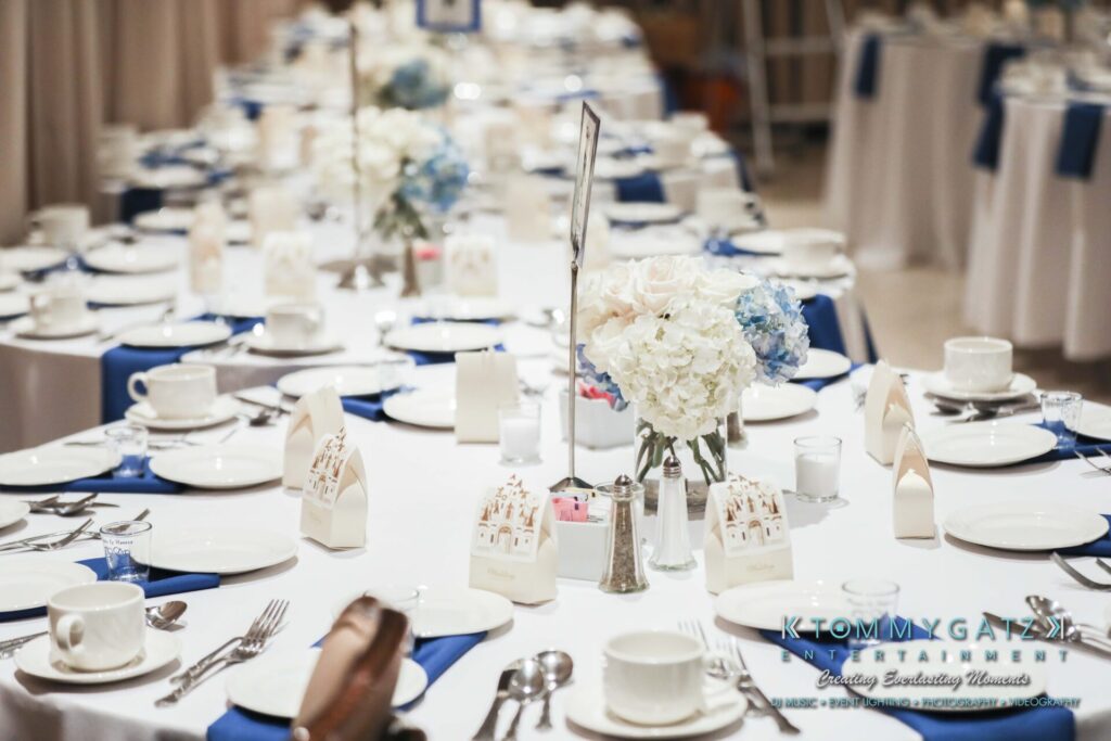 A beautifully set wedding reception table with white and blue accents, featuring plates, cutlery, and floral centerpieces.