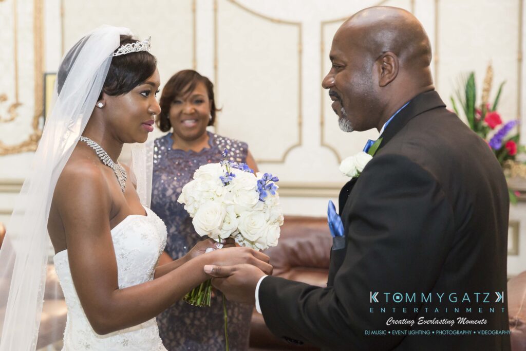 A bride and groom hold hands, facing each other, with a bouquet of white flowers. A smiling woman stands in the background, wearing a blue dress.