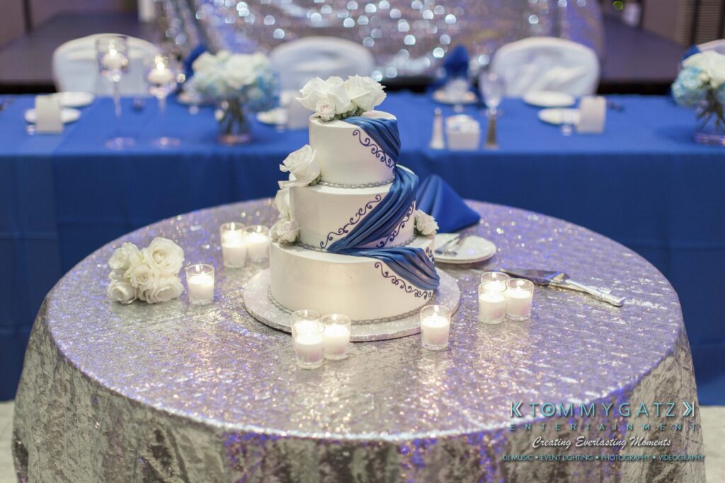 A three-tiered white wedding cake with blue accents on a round table, surrounded by small candles. Background features a blue and silver decorated table with floral arrangements.