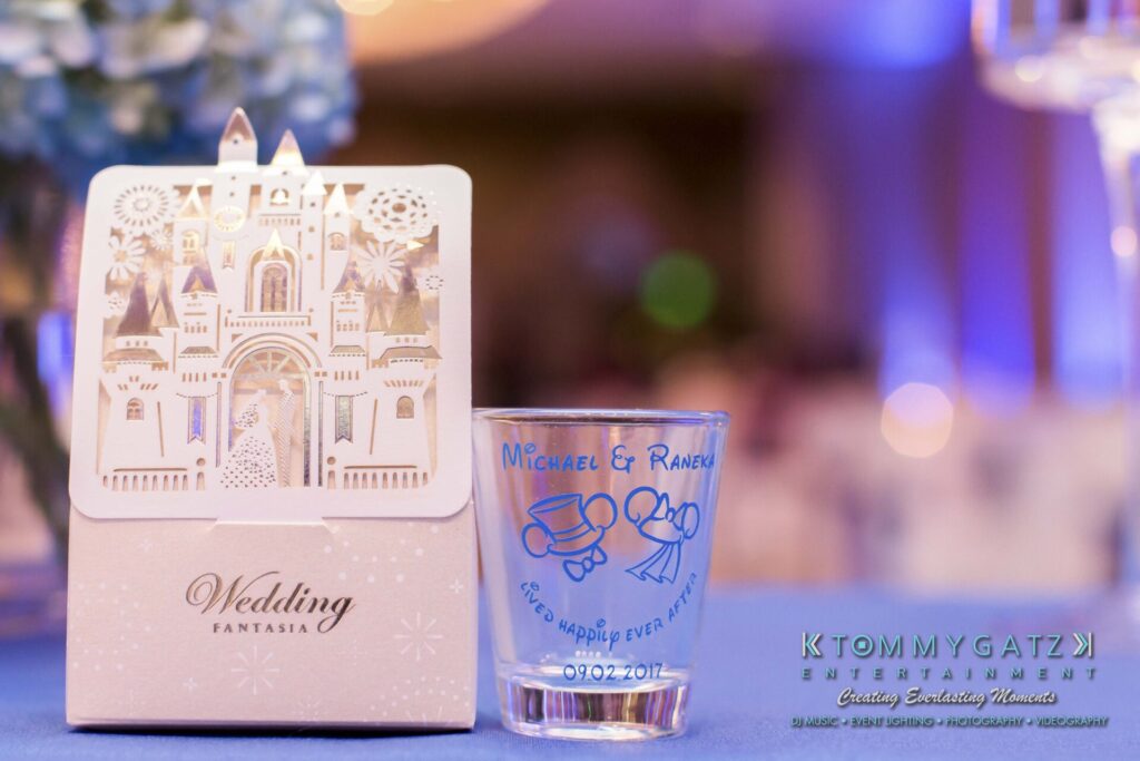 Wedding-themed decor with a card featuring a castle and a customized glass with "Michael & Raven" and wedding date, on a table. Background shows a blue-lit event setting.
