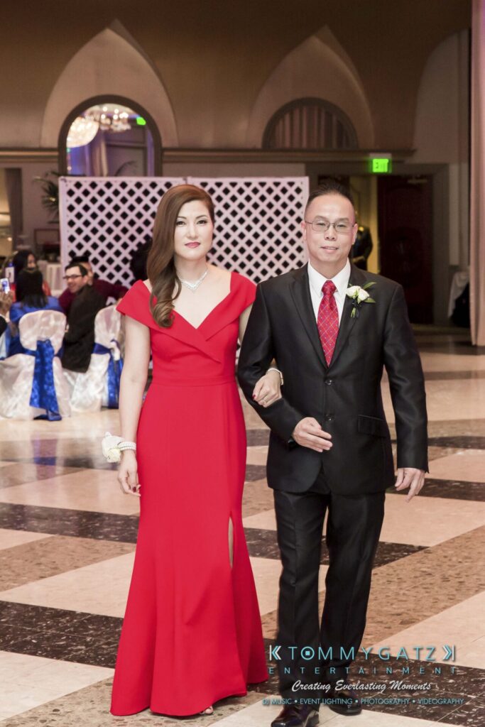 A woman in a red dress and a man in a black suit walk arm-in-arm in a decorated event hall.
