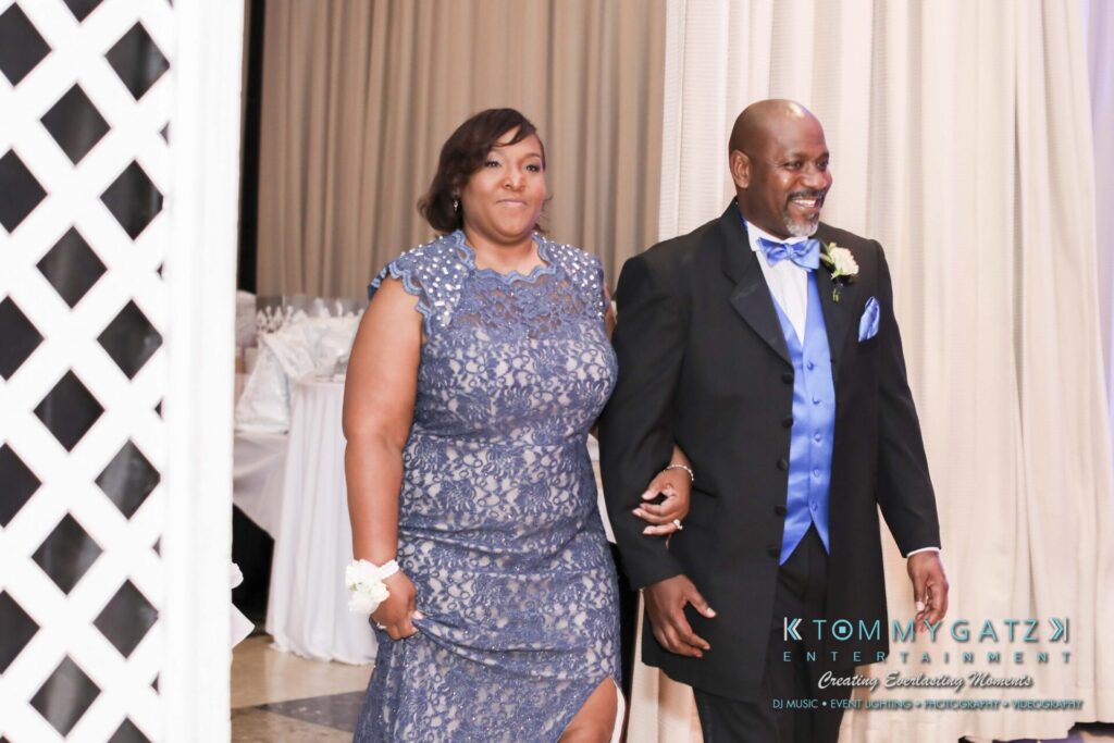 A man and woman in formal attire walk through a decorated venue. Both are smiling. The man wears a black suit with a blue vest and bow tie, and the woman is in a blue lace dress.