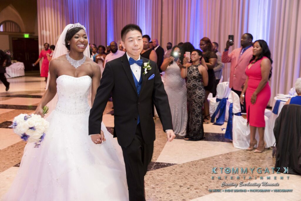Raneka-Michael-RECEPTION-Low-Res-WM-169 - TOMMY GATZ ENTERTAINMENT Bride and groom holding hands, smiling, walking into a reception hall with guests and photographers around them.