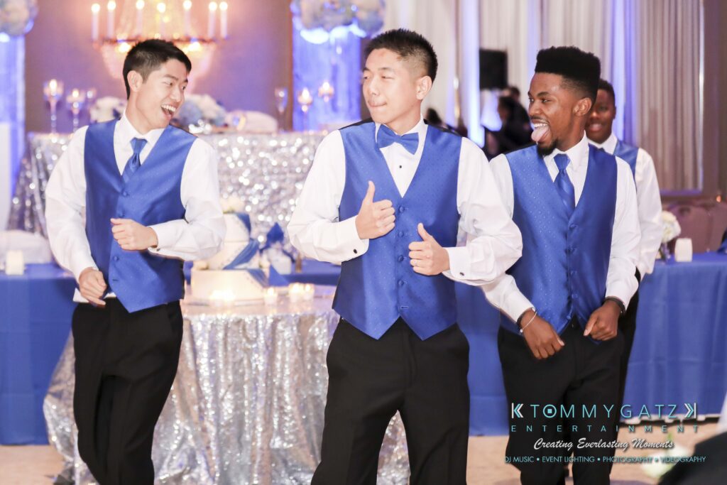 Three men in matching blue vests and black pants dance joyfully at an event, with silver and blue decorations in the background.