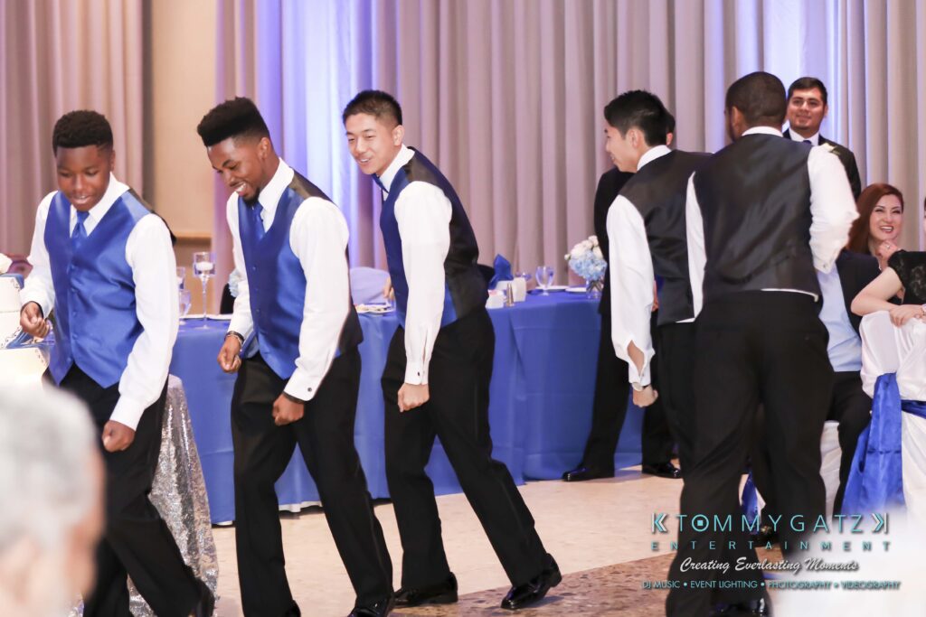 A group of young men in blue vests and black pants dance in formation at an event, while seated guests watch in the background.