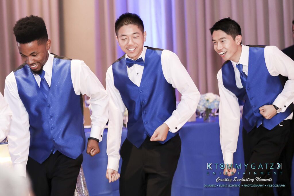 Three young men in blue vests and bow ties are laughing and moving energetically, likely dancing at an event.