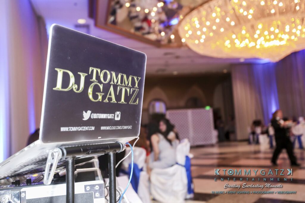 DJ setup with laptop displaying "DJ Tommy Gatz" logo in an elegant event hall. People are socializing in the background under large chandeliers.